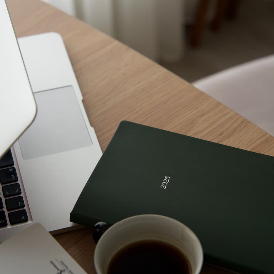 Laptop, en mörkgrön anteckningsbok och en kopp kaffe på ett skrivbord