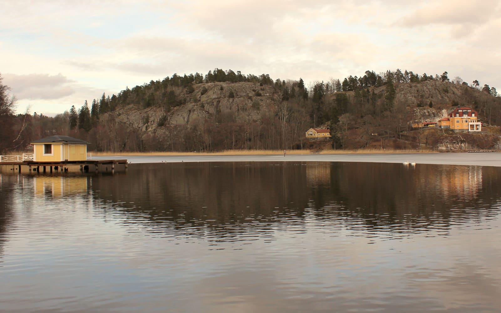 Tullingesjön en stilla dag – en gul paviljong på bryggan, skogsklädda berg och villor på andra stranden som speglar sig i vattnet